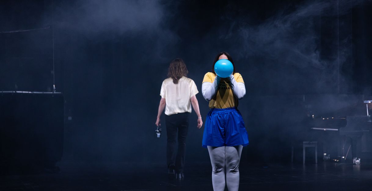 Excerpt from “My Kingdom for a Joke”: A person wearing silver leggings, blue sweatpants, and a yellow shirt holds a blue balloon in front of their face. In the background, a person dressed in black and white walks away.