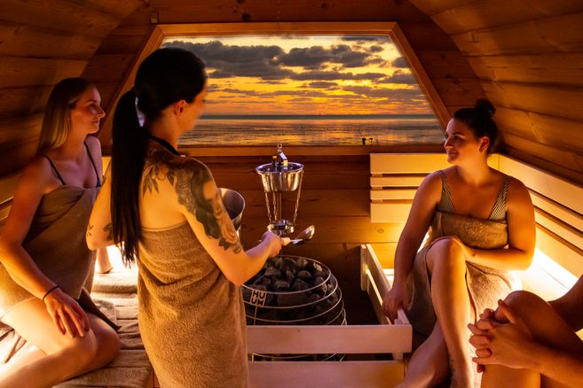 Drei Frauen sitzen in einer exklusiven Fass-Sauna mit Blick auf den Sonnenuntergang über dem Wasser.