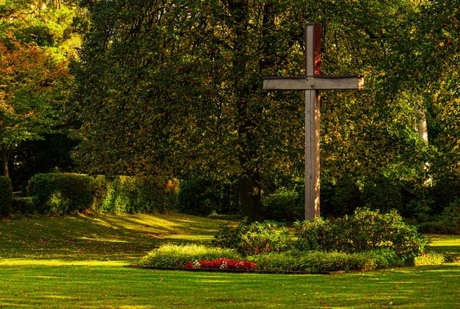 Ein großes Holzkreuz steht auf einem gepflegten Rasen, umgeben von Bäumen und Sträuchern im Sonnenlicht.