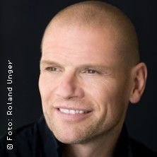 Porträt eines lächelnden Mannes mit Glatze vor schwarzem Hintergrund.