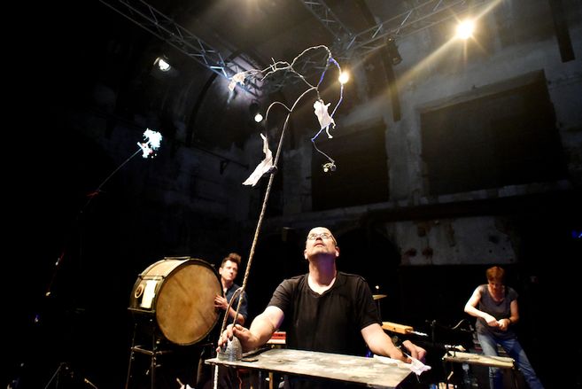 Ein Mann auf einer Bühne balanciert eine lange Stange mit Draht und Papier. Im Hintergrund sind Musiker und Instrumente zu sehen.