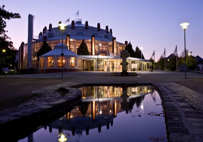 Ein beleuchtetes Theatergebäude bei Nacht mit Spiegelung im Wasser.