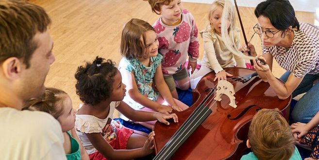Kinder sitzen um ein Cello, während eine erwachsene Person ihnen etwas zeigt.