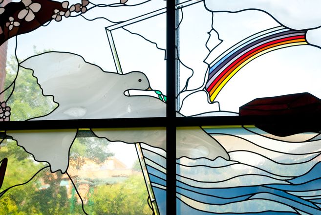 Buntes Kirchenfenster mit Taube, Regenbogen und stilisierten Wellen; im Hintergrund ist unscharf Grün zu sehen.