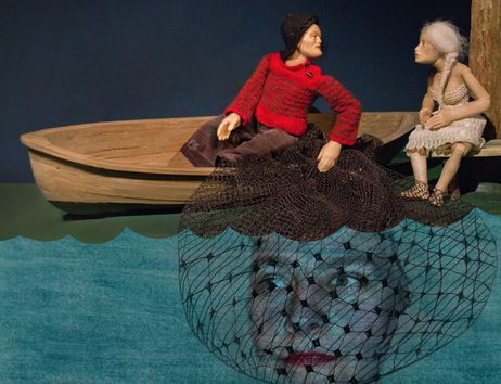 Zwei Puppen in einem Boot, eine in rotem Pullover, die andere in weißem Kleid. Darunter ein Gesicht hinter einem Netz im Wasser.