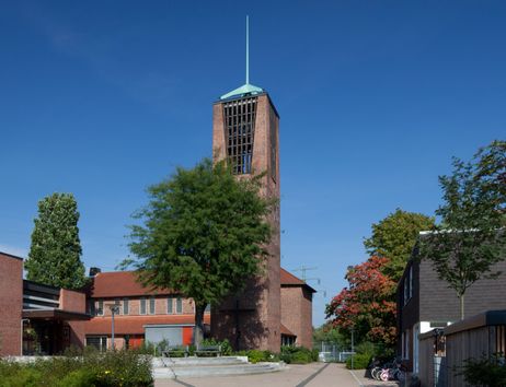 Eine Kirche mit einem hohen Turm und rotem Ziegeldach, umgeben von Bäumen und einem klaren blauen Himmel.