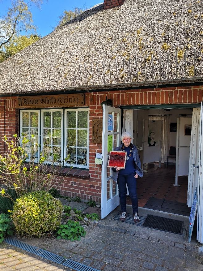 Eine Person steht vor einem Backsteinhaus mit Reetdach und hält ein Kunstwerk in der Hand. Über der Tür steht 'Kleine Waldschänke'.