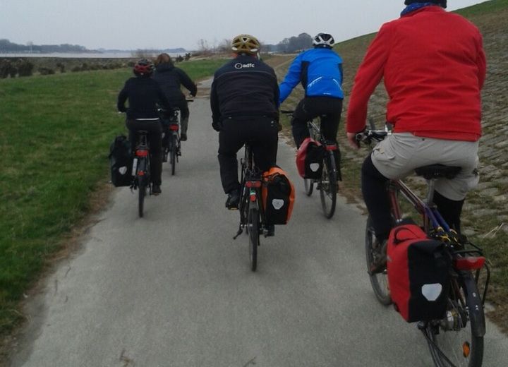 Fünf Radfahrer mit Gepäcktaschen fahren auf einem schmalen Weg entlang eines Deiches.
