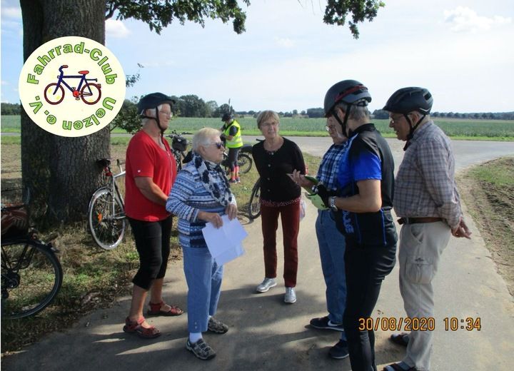Eine Gruppe von Menschen steht auf einem ländlichen Weg mit Fahrrädern im Hintergrund. Sie tragen Helme und diskutieren miteinander.