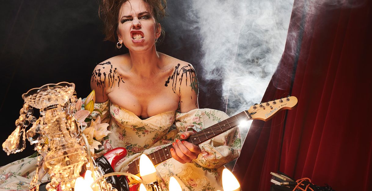 Emma Frankland, a female-presenting person with brown hair, stands in front of a curtain with an electric guitar and a rocking expression on her face.