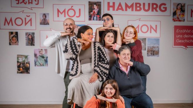 Eine Gruppe von Menschen posiert lächelnd vor einer Wand mit der Aufschrift 'Mein Hamburg' in verschiedenen Sprachen.