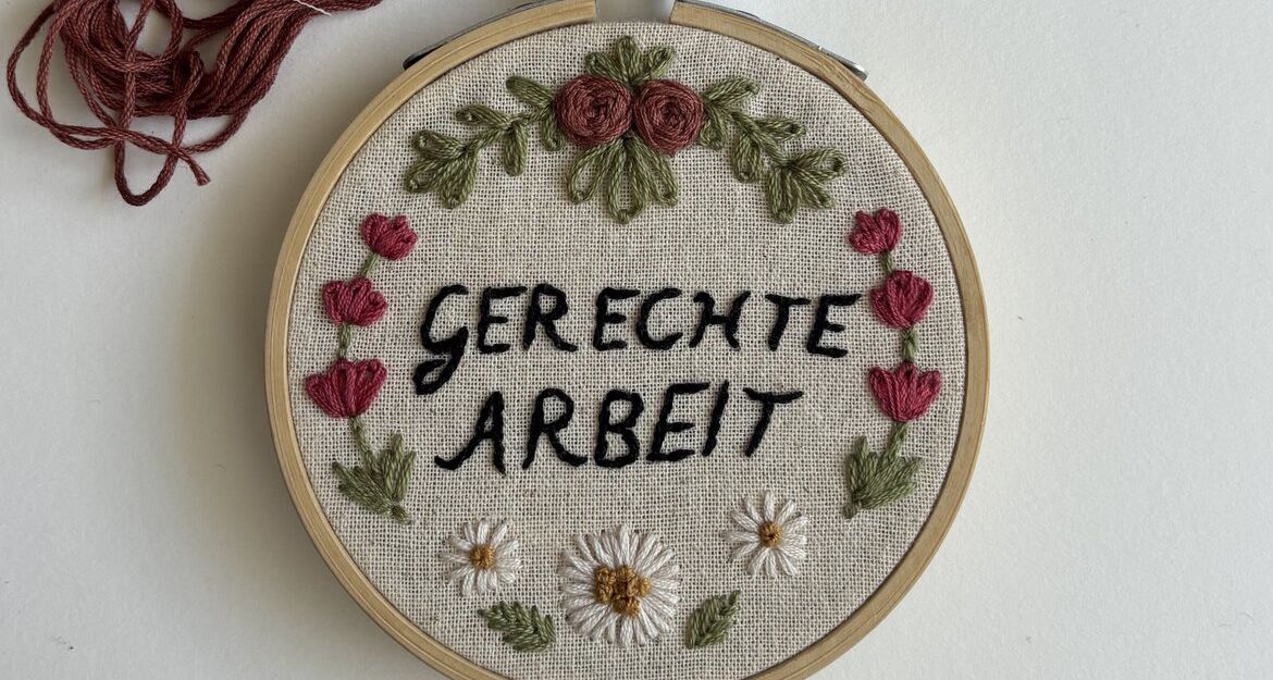 Ein Stickrahmen mit der Aufschrift 'Gerechte Arbeit', umgeben von gestickten Blumen und Blättern.