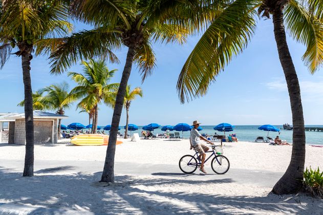 Das Bild zeigt den Strand an den Florida Keys, wo ein Mann zwischen Palmen Fahrrad fährt. 