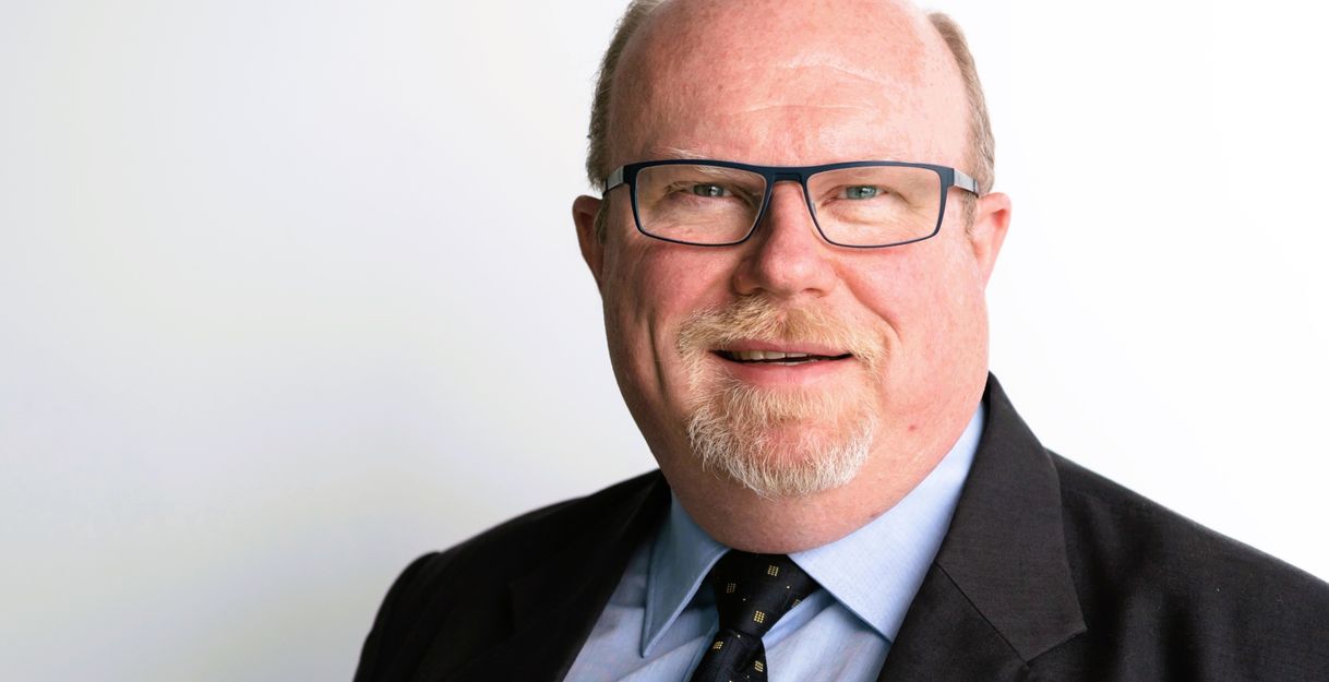 Porträt eines lächelnden Mannes mit Brille und Anzug vor weißem Hintergrund.