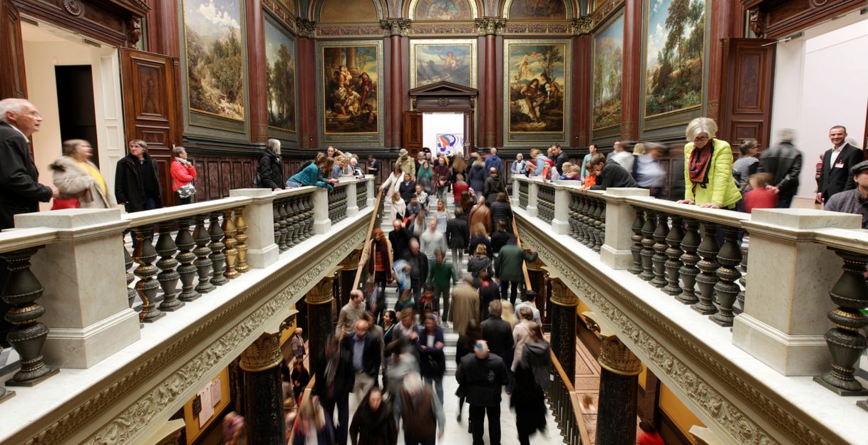 Menschenmenge in einem kunstvoll dekorierten Museum mit Gemälden an den Wänden.