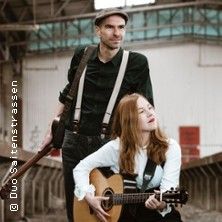 Ein Mann mit Hut und Hosenträgern steht neben einer Frau, die Gitarre spielt.