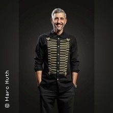 Ein Mann in schwarzem Hemd mit goldenen Verzierungen lächelt in die Kamera.