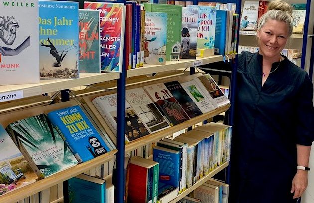 Eine Frau steht lächelnd neben Bücherregalen in einer Bücherei.