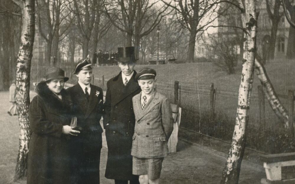 Schwarz-weiß Foto von vier Personen, die in einer parkänlichen Umgebung stehen. Von links: Frau mit feinem Mantel, junger Mann in Anzug, Mann mit Mantel und Zylinder, Junge mit Jackett und kurzen Hosen.