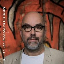 Porträt eines Mannes mit Brille vor einer bunten Wand.