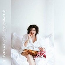 Eine Person sitzt in einem weißen Raum auf einem Bett und isst Pizza.