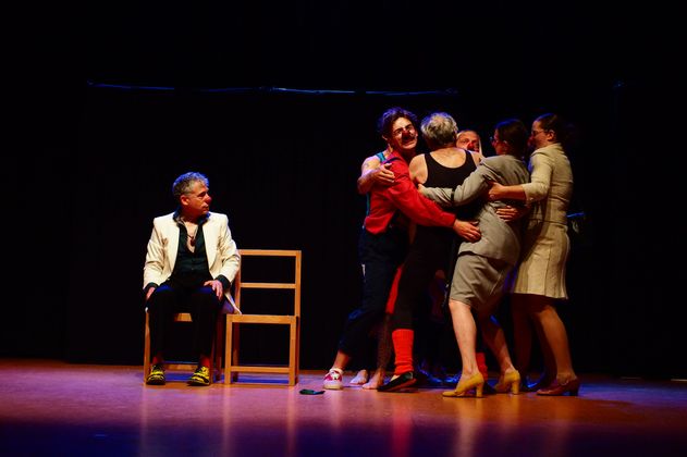 Eine Gruppe von Menschen auf einer Bühne, eine Person sitzt auf einem Stuhl, während andere sich umarmen.