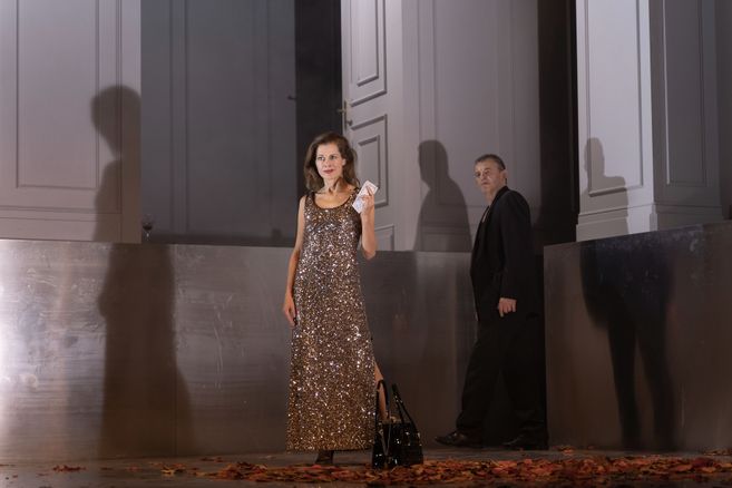 Frau in glitzerndem Abendkleid steht auf einer Bühne vor einer offenen Tür, daneben ein Mann in dunkler Kleidung; Herbstblätter liegen am Boden.