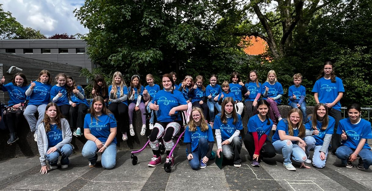 Eine Gruppe von Kindern in blauen T-Shirts sitzt und kniet draußen auf einem Platz. Sie lächeln und zeigen Daumen hoch.