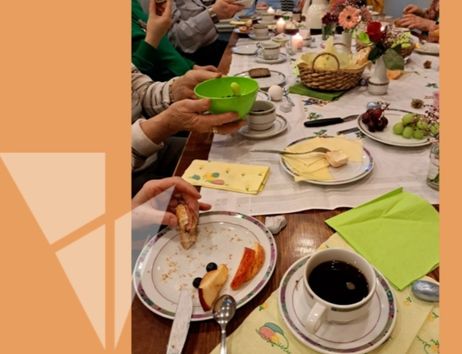 Menschen sitzen an einem gedeckten Frühstückstisch mit Kaffee, Obst und Gebäck.