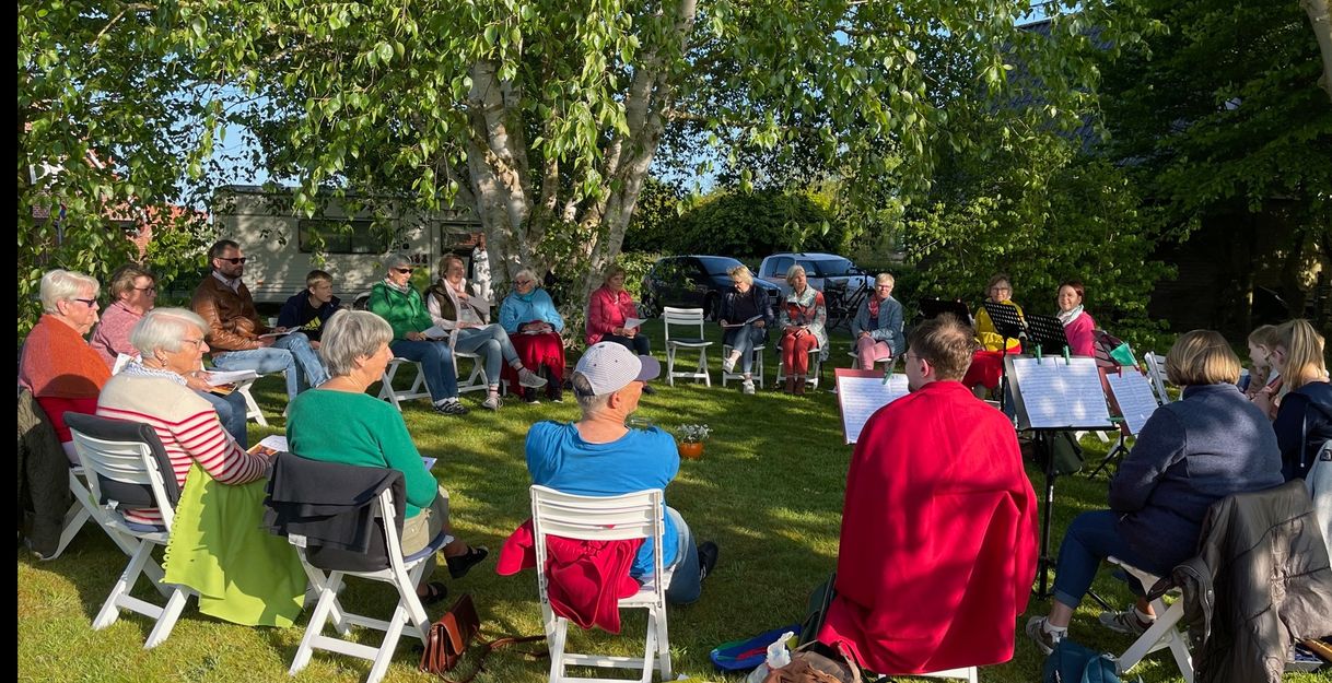 Eine Gruppe von Menschen sitzt im Kreis auf weißen Stühlen im Freien unter einem Baum.