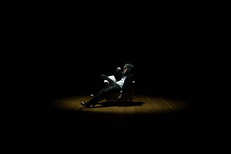 Person sitzt in einem Sessel im Dunkeln, beleuchtet von einem Spotlicht.