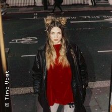 Eine Frau mit langen Haaren und einem roten Oberteil steht auf einer Straße. Sie trägt eine schwarze Jacke und hat zwei Haarbündel auf dem Kopf.