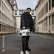 Person mit Boombox steht in einer leeren Straße zwischen hohen Gebäuden.