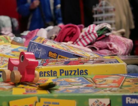 Kinderflohmarkt mit Spielzeug, Puzzles und Kleidung; im Vordergrund ein rotes Bobbycar aus Holz, im Hintergrund unscharf stehende Personen.
