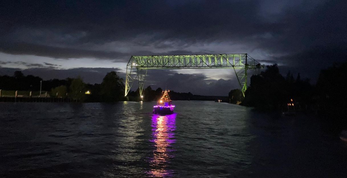 Ein beleuchtetes Boot fährt nachts unter einer Brücke hindurch, die in grünem Licht erstrahlt.
