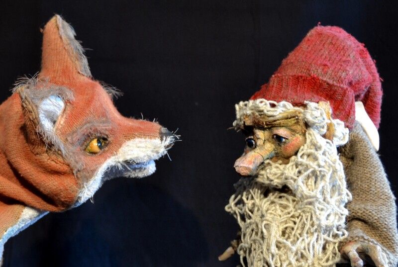 Eine Puppe eines Fuchses und eine Puppe eines alten Mannes mit roter Mütze stehen sich gegenüber.