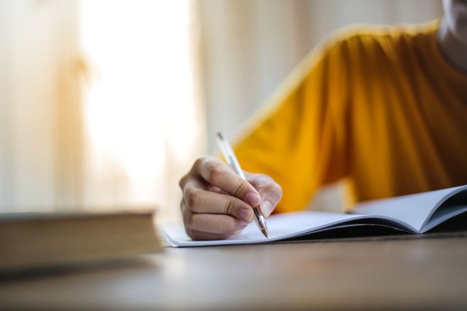 Person in gelbem Shirt schreibt mit einem Stift in ein aufgeschlagenes Notizbuch am Tisch, links liegt ein Buch im Vordergrund.