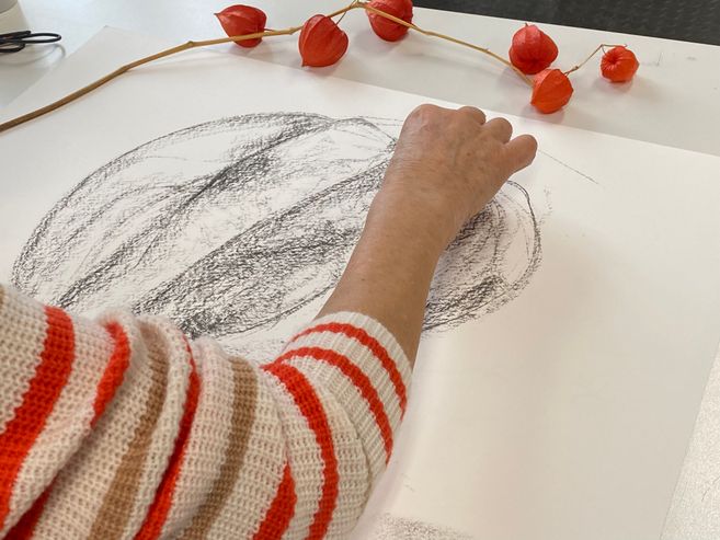 eine Frau in einem rot-weiß geringeltem Pullover zeichnet mit Kohle vergrößert einen Stengel mit orangenen Lampionblumen 