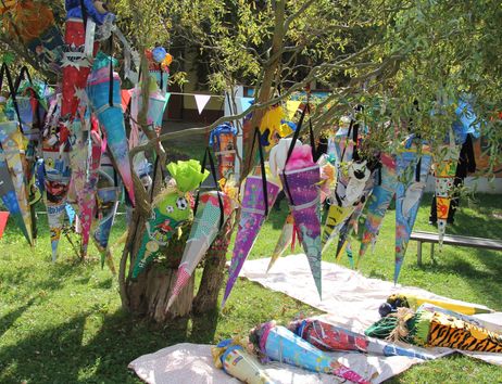 Ein Baum, an dem bunte Schultüten hängen, steht auf einer Wiese. Einige Schultüten liegen auf Decken am Boden.