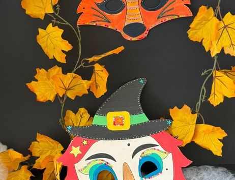 Zwei bunte Masken, eine Fuchsmaske und eine Hexenmaske, hängen an einer Wand mit Herbstblättern und Spinnweben.