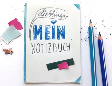 Ein Notizbuch mit der Aufschrift 'Lieblings-Mein Notizbuch' liegt neben zwei blauen Buntstiften und bunten Papierschnipseln.