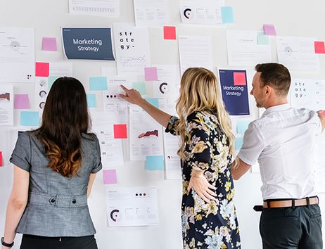 Es sind drei Personen zu sehen, die vor einer Wand mit Post-Its stehen