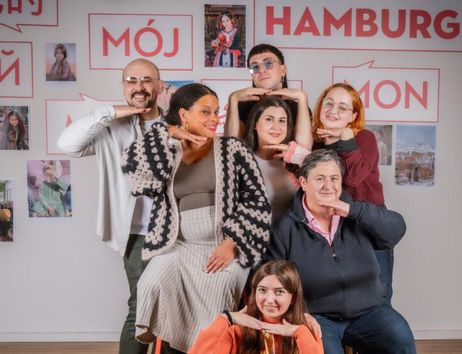 Eine Gruppe von Menschen posiert lächelnd vor einer Wand mit der Aufschrift 'Mein Hamburg' in verschiedenen Sprachen.