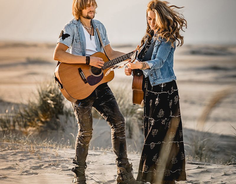 Zwei Personen spielen Gitarre in einer sandigen Dünenlandschaft.