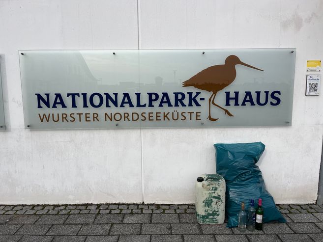 Schild des Nationalpark-Hauses Wurster Nordseeküste mit Kiebitz-Silhouette; davor stehen ein Müllsack, ein Kanister und mehrere Flaschen.