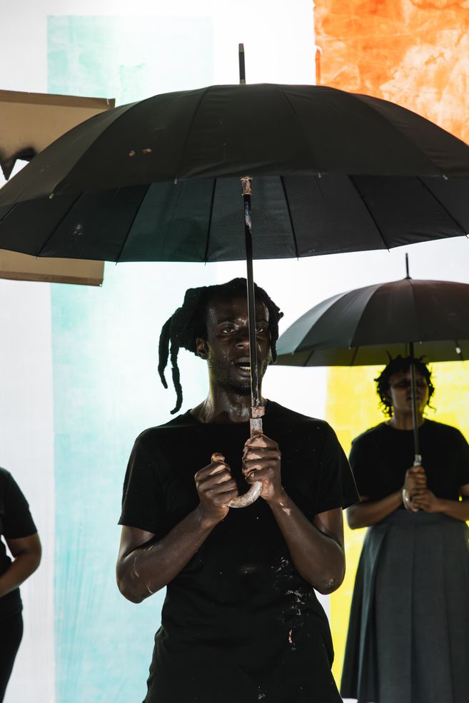 Ausschnitt aus der Produktion DAMBUDZO von nora chipaumire. Eine Person steht mit einem Regenschirm vor einer bunten Wand.