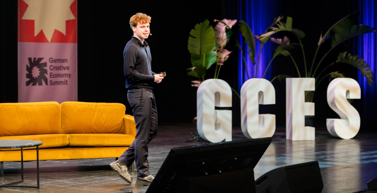 Excerpt from the German Creative Economy Summit. A stage with a speaker can be seen. In the background, there is a couch and the letters GCES.