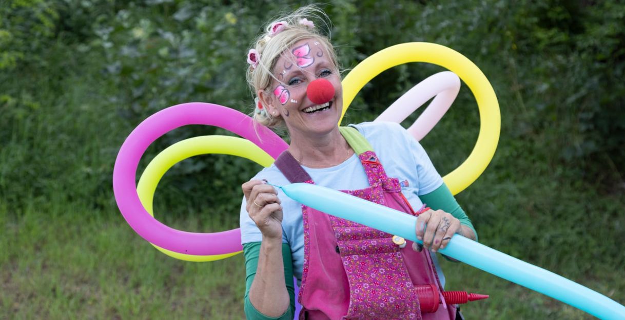 Eine fröhliche Person mit Clownsnase und bemaltem Gesicht hält bunte Luftballons in einem Garten.