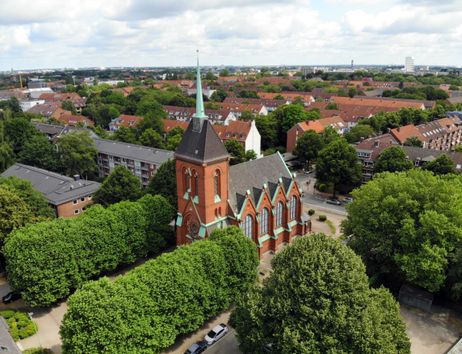 Luftaufnahme einer roten Backsteinkirche mit grünem Dach inmitten einer Wohngegend mit vielen Bäumen.