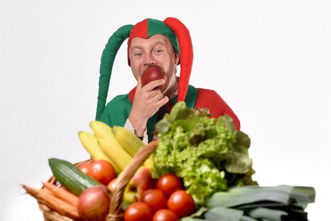 Ein Mann in einem bunten Narrenkostüm isst einen Apfel hinter einem Korb voller Obst und Gemüse.